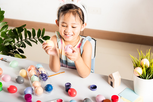 Happy Asian little girl kid using brush painting egg at home, Children after paint Easter eggs already with colors on table she playing with eggs, Happy Easter day concept, Preparing DIY handmade