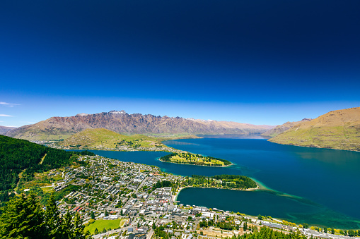 This November 2021 daylight image shows Tāhuna Queenstown, Aotearoa New Zealand. The turqouise waters of Lake Wakatipu dominate the scene with the city centre along its shores. In the distance are Tākitimu the Remarkables mountain range with a hint of snow. This image was taken on a sunny, spring day with clear skies from near Ben Lomond Scenic Reserve. Watercraft have been removed from this image.