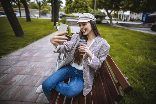Photo of young woman using a smart phone, sitting on the bench in the park, smiling and takes selfie by her phone.