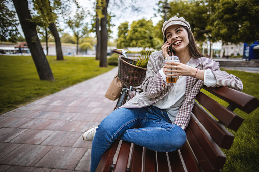 Photo of young woman using a smart phone, sitting on the bench in the park, smiling.