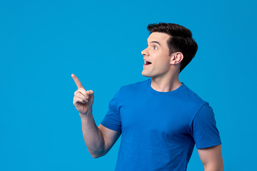 Excited young Caucasian man looking sideways and pointing up in light blue isolated background