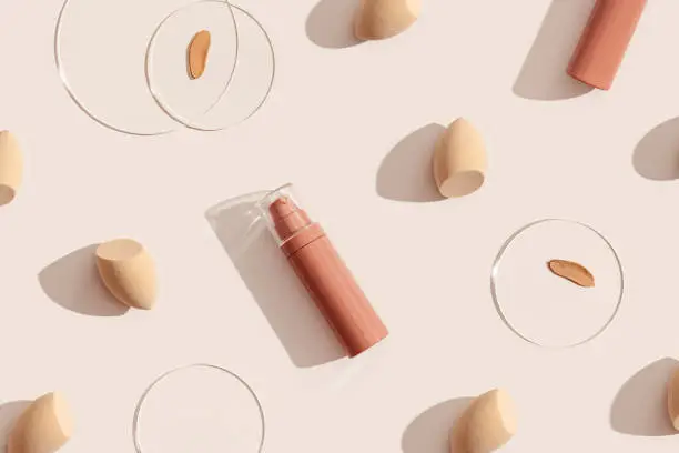Photo of Bottle of makeup foundation cream, samples on glass disk, beauty blender sponge on beige color background with hard light. Skin care beauty product. Top view package concealer base container