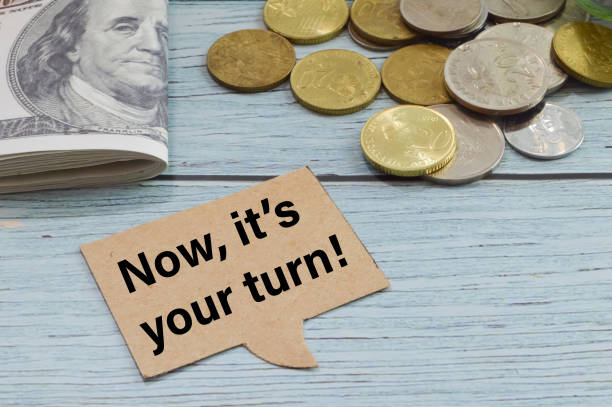 istock Speech bubble note written with NOW IT'S YOUR TURN with coins and money banknotes. 1397468781