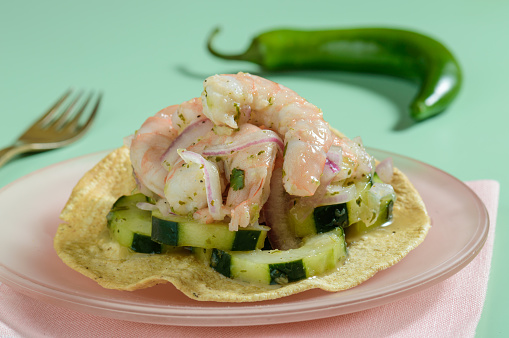 Aguachile. Mexican take on ceviche. Delicious and spicy marinated seafood dish with shrimp, serrano chili, cucumber, lime juice and red onion. With pink and green background.