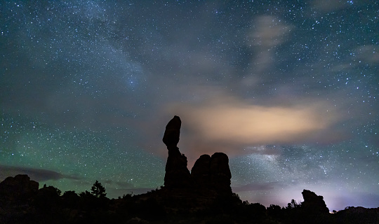 Iconic Balanced Rock silhouetted against a mixed sky of cloud and the Milky Way in Arches National Park, Utah.
