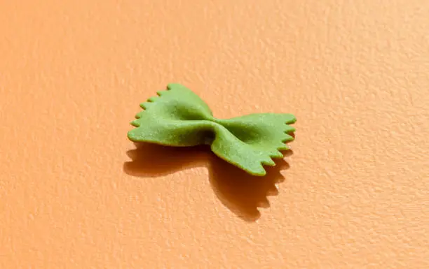 Single ribbon-shaped pasta isolated on orange background in bright light. Spinach pasta in the shape of a bow tie, minimalist on a colorful table.
