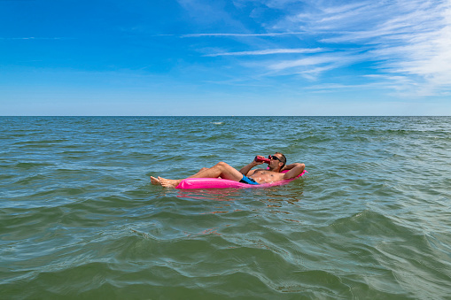 Tanned man relax on pink inflatable mattress at sea. Successful Guy in sunglasses enjoying vacation and cool drinks