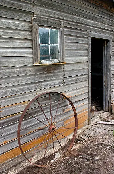 A rusty wagonwheel sits outside a brokedown barn.