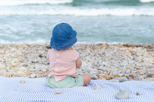 Back view of a baby girl sitting on a blue striped towel at a pebble beach at the end of summer.