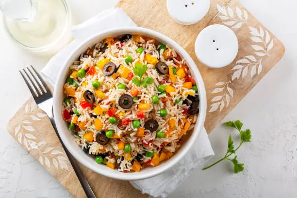 Photo of Rice Salad with fresh vegetables, bell bebber, tomato, green pea, black olives and olive oil,  on a white table. Directly above.