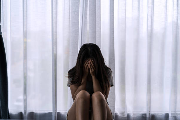 暗い寝室に座って落ち込んでストレスを感じている孤独な若い女性、否定的な感情とメンタルヘルスの概念 - ストレス ストックフォトと画像
