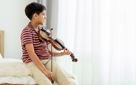 Indian handsome teenage boy, playing, practicing violin musical instrument with happiness in bedroom at cozy home in leisure time, smiling, having copy space. Education, Lifestyle, Creativity Concept