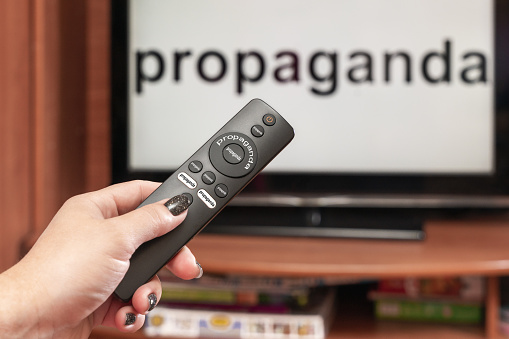 A woman's hand holds the TV remote control, all the buttons on the remote control are signed propaganda, on the TV there is an inscription propaganda.