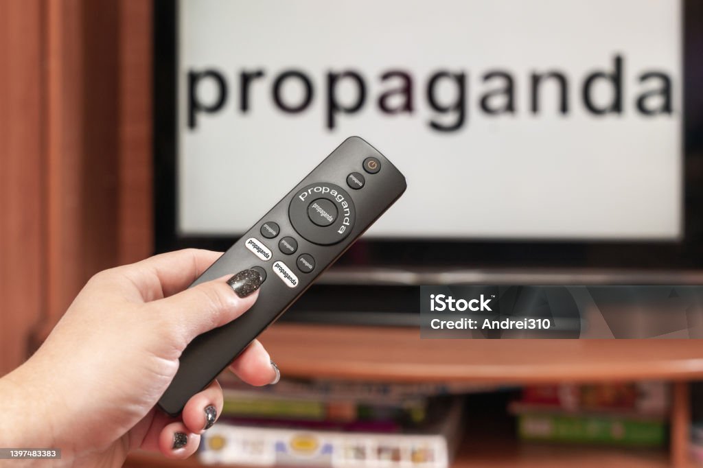 A woman's hand holds the TV remote control, all the buttons on the remote control are signed propaganda, on the TV there is an inscription propaganda A woman's hand holds the TV remote control, all the buttons on the remote control are signed propaganda, on the TV there is an inscription propaganda. Arguing Stock Photo