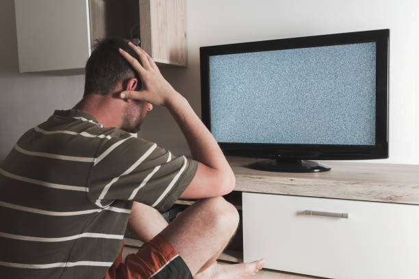 Young caucasian man sitting with head in hands looking to grey graining television. Depression, psychic, mentally ill concept stock photo