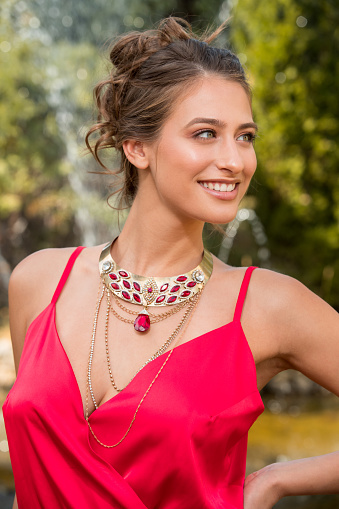 Beautiful young woman in red sleeveless top wearing ruby jewelry, standing in public park and smiling looking away, beauty and fashion industry