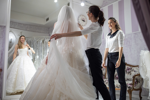 Caucasian female customer, a future bride, at the bridal shop trying on wedding dress, with a help of saleswoman