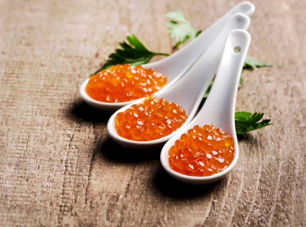 primer plano de caviar rojo de lujo en cuchara blanca con perejil sobre fondo de mesa de madera. - desovar fotografías e imágenes de stock