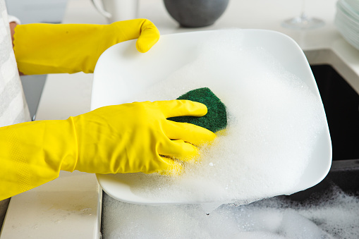 Unrecognizable hand with yellow cleaning glove scrubbing dishes close up in kitchen.
