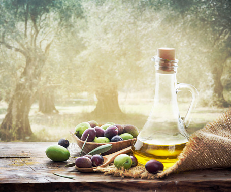 Olives and bottle of Extra virgin olive oil on wooden table in olive garden. Traditional homemade olive oil and olive trees with sunlight in background.