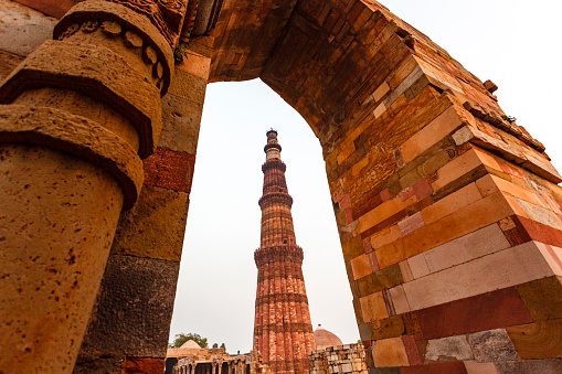 This image features the Qutub Minar, one of Delhi's most iconic landmarks, shrouded in the soft haze of an early morning. Standing at a height of 73 metres, the Qutub Minar is the tallest brick minaret in the world and is known for its intricate carvings and inscriptions. The haze adds an ethereal quality to the photograph, slightly obscuring the minaret's details but enhancing its mystique. This atmospheric condition offers a different perspective on a well-known monument, adding a layer of complexity and mood. The image aims to capture both the historical significance and the natural surroundings of the Qutub Minar, presenting it as a must-see attraction with ever-changing moods and appearances.