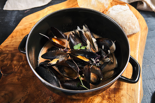 Mussel stew pot with creamy sauce, baguette on stone table. Mussels soup pot contemporary style. Aesthetic composition with seafood. Gastronomy mussel pot dinner concept. Stone table and wooden tray
