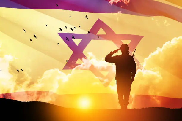 Photo of Silhouette of soldier saluting against the sunrise in the desert and Israel flag.