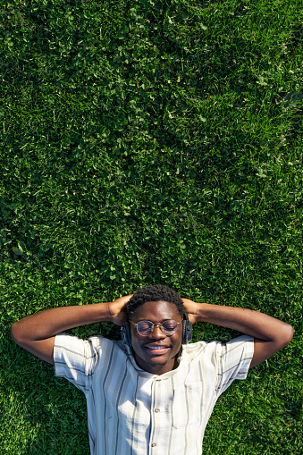Top view of black man relaxing. Happy black male lying on grass listening to music with headphones. Copy space Vertical image. Relaxation concept.