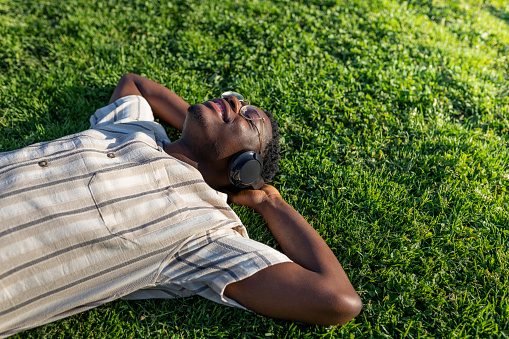 Black man relaxing outdoors. Happy black male lying on grass listening to music with headphones. Copy space. Relaxation concept.