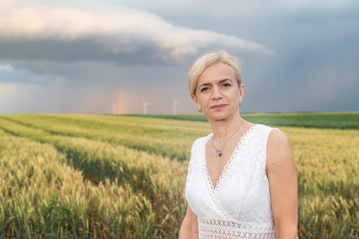 Woman in white sleeveless dress standing in agricultural field with wind turbines under dramatic sky