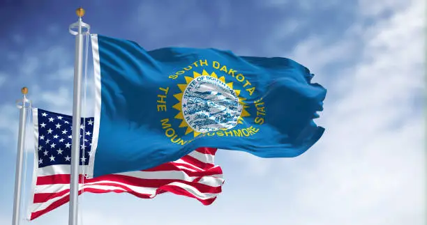 The South Dakota state flag waving along with the national flag of the United States of America. South Dakota is a U.S. state in the North Central region of the United States