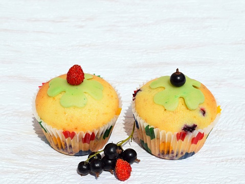 Summer baking - delicious homemade muffins with berries in colorful paper cups, decorated with marzipan cloverleaf, fresh black currant and strawberry. Creativity, originality, romantic baking