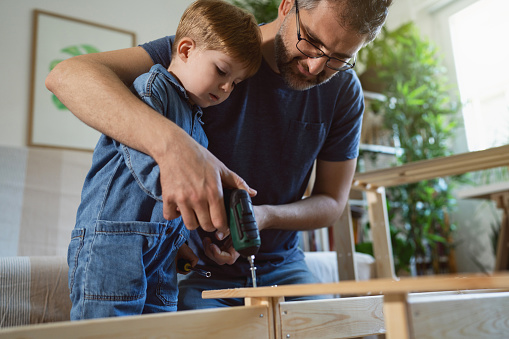 Caucasian toddler boy assist to his father, while he assembling new furniture at home