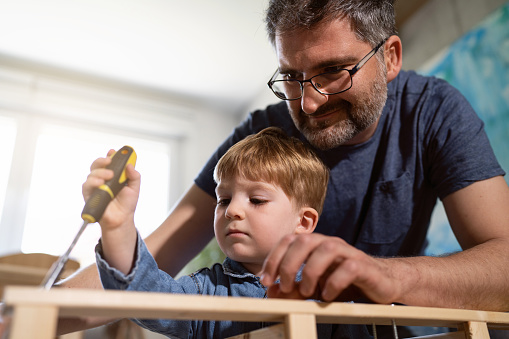 Curious Caucasian toddler boy assist to his father, while he assembling new furniture at home