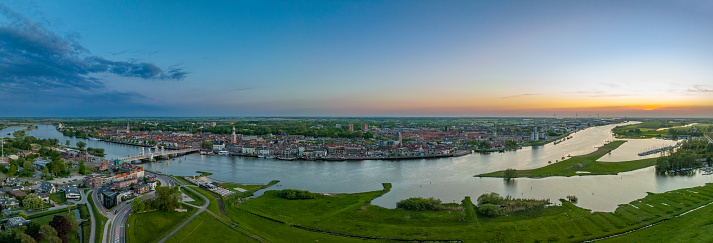 Panoramic aerial view on the Hanseatic League city Kampen at the river IJssel during a springtime sunset in Overijssel. The flow of the river is leading towards the setting sun in the distance while lights are popping up in the city at the end of a beautiful springtime day.
