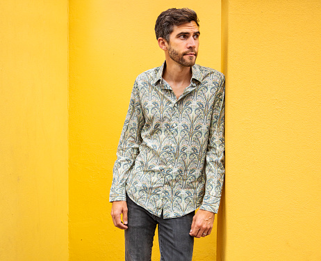 Stylish young man with a beard wearing a paisley shirt leaning against a bright yellow wall outdoors