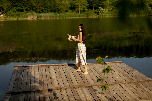 Happy young woman standing on the wooden pier at the calm lake on a hot summer day
