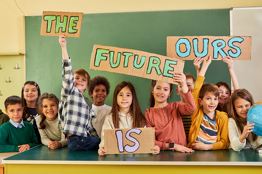 Happy elementary students holding placards in the classroom with a message that the future is theirs and looking at camera.