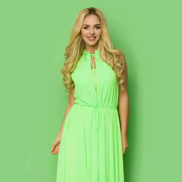 Beautiful smiling blond woman in a neon green maxi dress. Three quarter length studio shot against green background.
