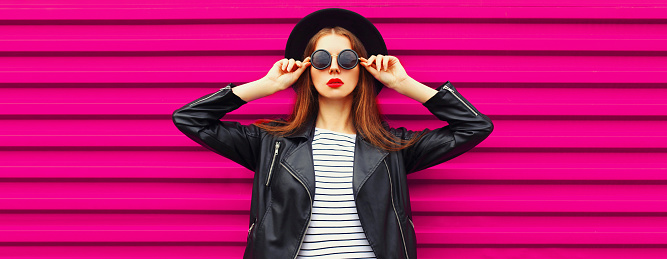 Fashionable portrait of stylish woman model posing wearing black rock biker jacket and round hat on pink background, blank copy space for advertising text