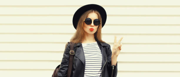 Fashionable portrait of stylish young woman blowing her lips and posing wearing black rock biker jacket, round hat and backpack on white background, blank copy space for advertising text stock photo