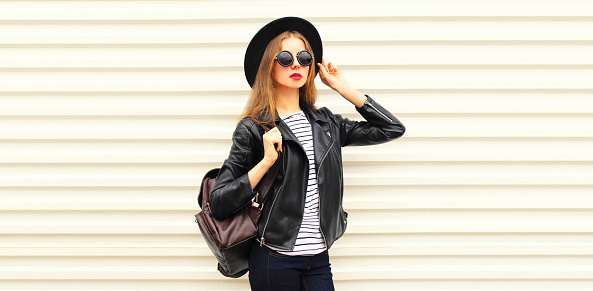 Fashionable portrait of stylish young woman posing wearing black rock biker jacket, round hat and backpack on white background