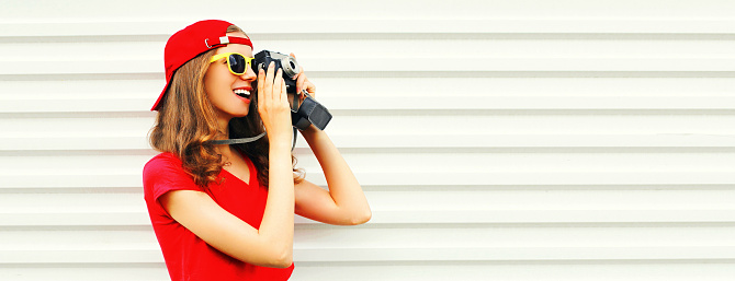 Portrait of happy smiling young woman photographer taking picture on film camera wearing red baseball cap on white background, blank copy space for advertising text