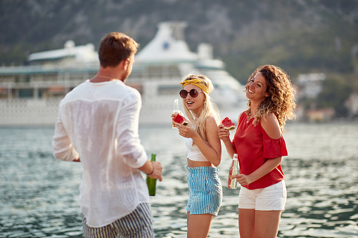 A group of cheerful friends having drink and eating watermelon on the dock on a beautiful day on the seaside. Friendship, holiday, sea