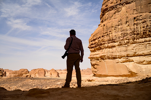 Full length rear view of landscape photographer standing in shade with camera over shoulder, looking at Jabal Alfil and neighboring rock formations.