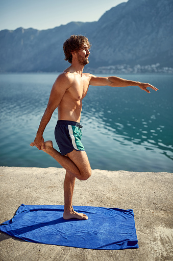 Man doing stretching exercise on jetty by water. Beautiful background of sea and mountains. Sport, vitality, recreation concept.