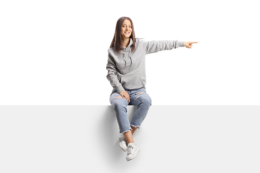 Female teen sitting on a blank board and pointing to the side isolated on white background