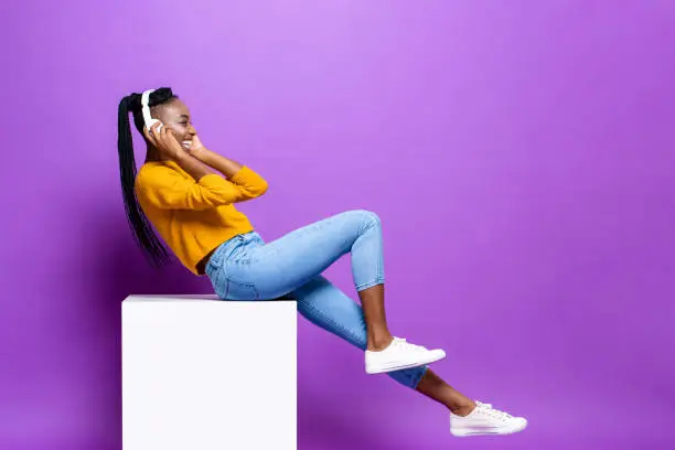 Photo of Smiling happy young African-American woman wearing headphone sitting on stool listening to music in studio purple color isolated background