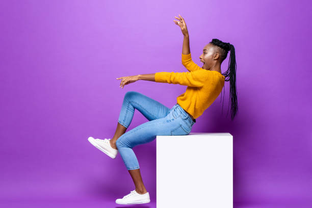 Fun excited young African-American woman sitting on stool raising leg and hands in studio purple color isolated background Fun excited young African-American woman sitting on stool raising leg and hands in studio purple color isolated background person falling backwards stock pictures, royalty-free photos & images