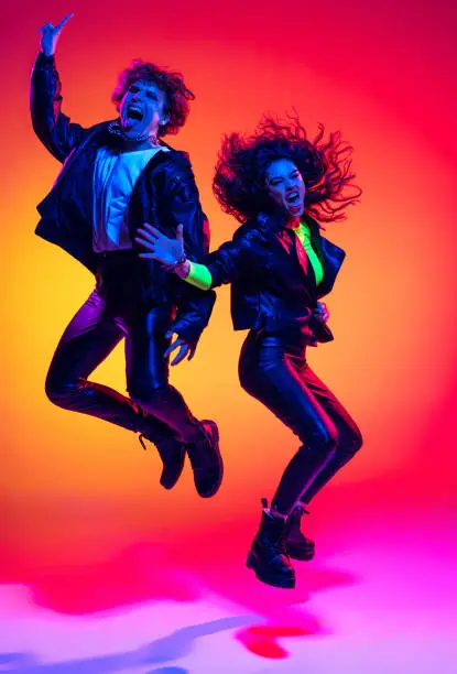 Excited young man and girl in black leather outfits having fun, gesturing, shouting and jumping on yellow-red background in neon light. Concept of style, music, fashion and youth